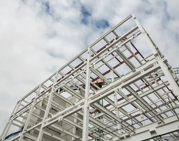 Structură metalică albă a unei clădiri în montaj, văzută de jos, pe cer cu nori